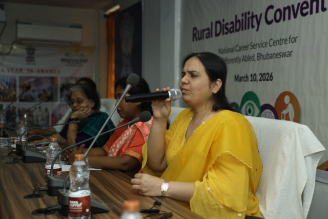 close-up of Nidhi Goyal speaking at a panel. She is holding a mic. She is wearing a yellow suit. The other speakers can be seen in the background, as well as a banner behind her.