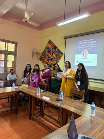 A group photo with Nidhi Goyal and the organisers. She is holding a certificate and smiling at the camera. Behind them is a projector with the details of the event on it - "Parwaaz 2026, Many Lives, Many Experiences: Disability and Intersectionality, Speaker Session: Nidhi Goyal" with the date, time, and venue at the bottom.