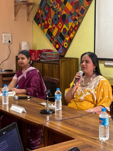 Close-up of Nidhi Goyal sitting at a table and speaking into a mic. She is wearing a yellow kurti with white patterned details and a necklace. There is another woman sitting next to her, listening.