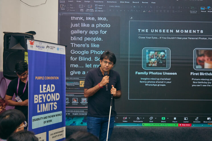 Sourabh speaking into the microphone, standing in front of his presentation and a zoom screen showing captions.