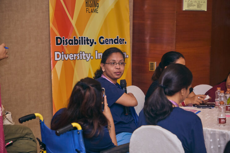 Close-up of one of the participants sitting around a table. There is a Rising Flame standee against a wall behind her. Next to her is someone on a wheelchair turned away from the camera. 