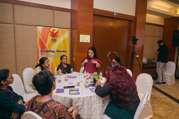Wide shot of one of the round tables. One of the speakers is a little person who is standing and speaking into a mic while the others listen. 