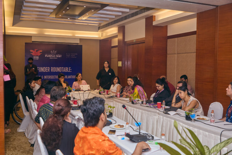 Wide shot of the roundtable in a conference room. At the far end at the head of the table are Raj Mariwala and Nidhi Ashok Goyal, sitting in front of the event banner. On either side of them are funders and disabled founders with conference mics on the table. There is also an ISL interpreter on Nidhi's left. 