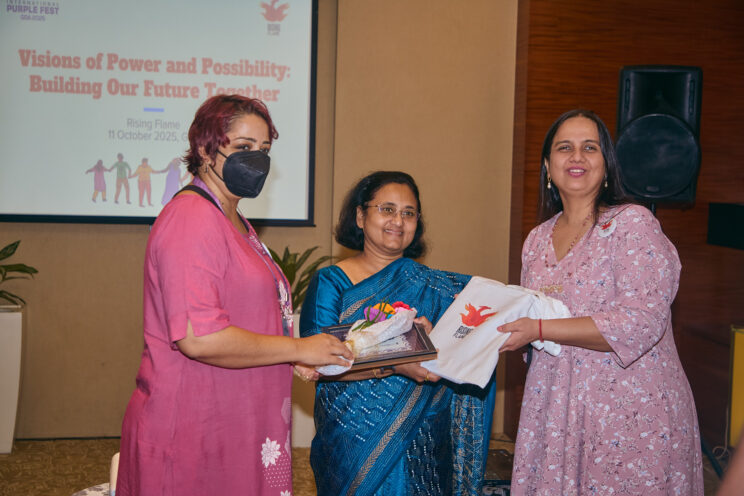 Smt. Ipsita Mitra posing with a Rising Flame tote bag and momento. She is standing in between Srinidhi Raghavan and Nidhi Goyal. 