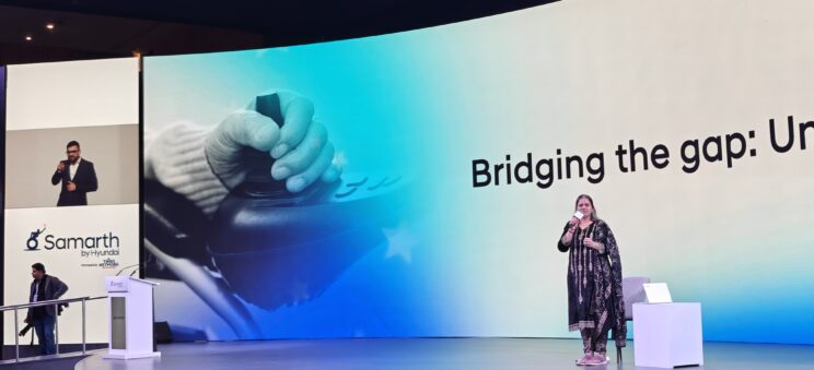 Nidhi Goyal on a stage in front of a large LED backdrop which reads, "Bridging the Gap." She is standing near a small table with a laptop. She is wearing a black salwar kameez with gold details. She is speaking into a mic. There is also a Sign Language interpreter on a screen off to the side.