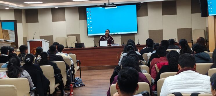 Full shot of a room from the back. Nidhi is in the front, sitting at a desk with her laptop in front of a projector. There are students listening to her or taking notes in their seats.