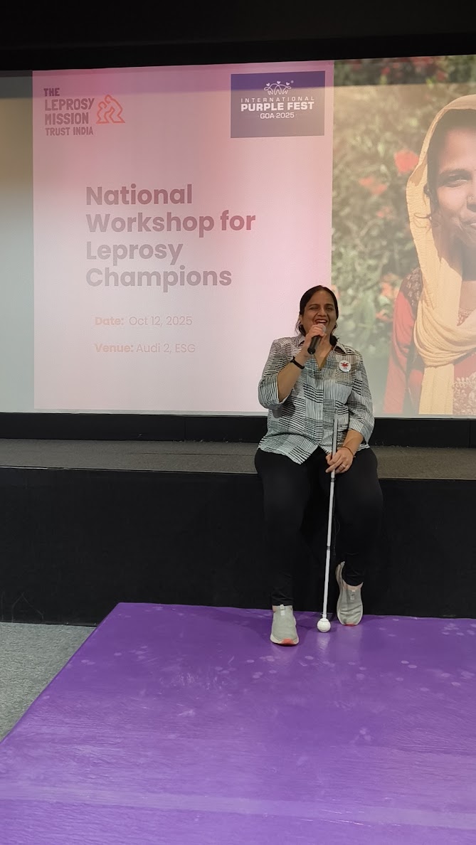 Nidhi sitting on the edge of a small stage. she is in front of projector with the session name on it - National Workshop for Leprosy Champions. She is speaking into a mic.