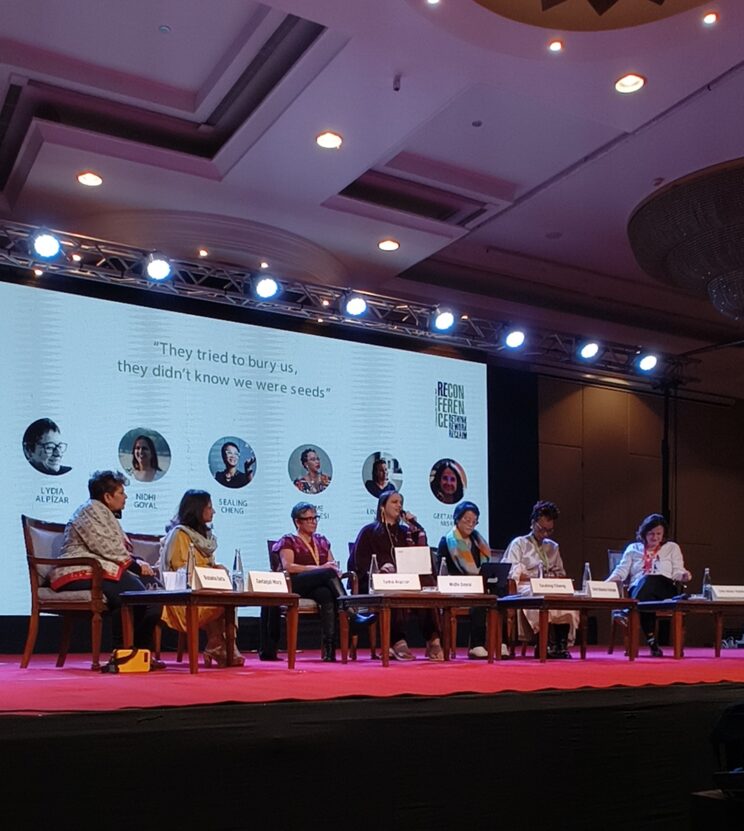Nidhi on the panel, she is speaking while the others look at her. LED screen behind has the name of the plenary and photos of the panellists.