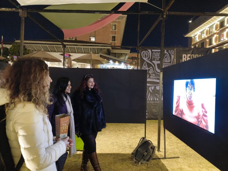 3 women standing in front of a TV showing the 'Still We Rise' video - The frame is of Rusha Chowdhury speaking. The women are smiling at the screen.