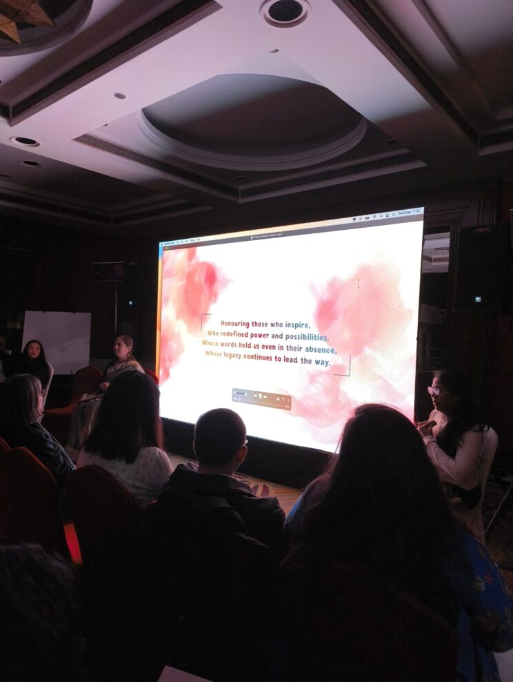 In a dark room, people are watching a video on an LED screen. On it is text that reads, "Honoring those who inspire, Who redefined power and possibilities, Whose words hold us even in their absence, Whose legacy continues to lead the way."