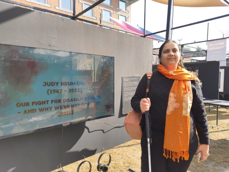 Nidhi in a black sweater and orange scarf, standing and smiling next to the TV playing the video. It is on Judy Heumann's title card - "Judy Heumann (1947-2023) Our fight for disability rights - and why we're not done yet (2016)"