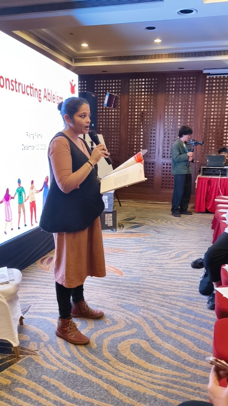 Prathama Raghavan standing at the front of a room with people in the audience. She is speaking into a mic, holding a copy of Holding Space Part 1.