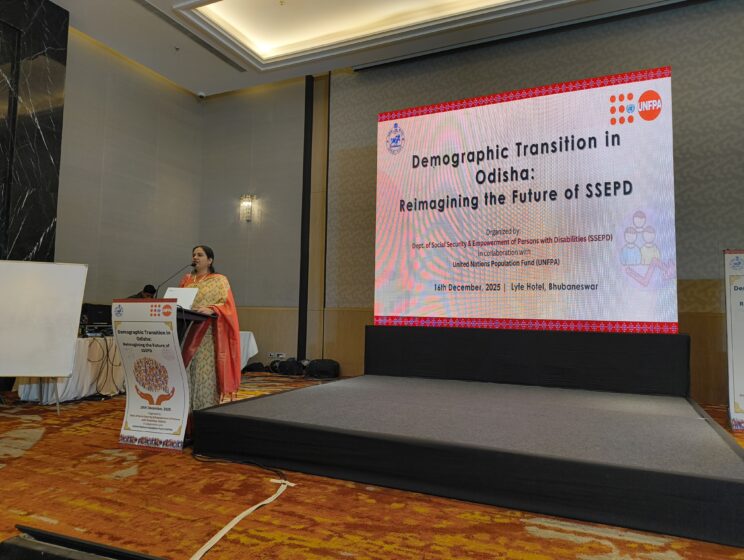 Nidhi is speaking at a podium with a mic in her hand. She is standing in front of a large projector with the session details on it. She is wearing a light coloured saree with peach accents.