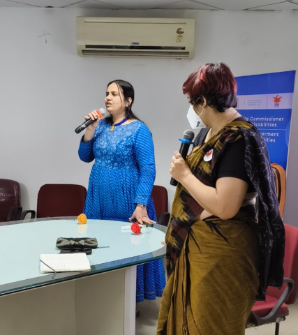 Nidhi Goyal and Srinidhi Raghavan standing at the head of a conference table. Nidhi is speaking into a mic. She is wearing a blue long sleeved kurta/dress. Srinidhi is wearing a black and gold saree and a white face mask.