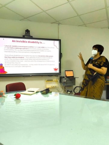 Srinidhi Raghavan standing in front of a TV projecting a presentation showing what an invisible disability is. She has a white face mask on and she is wearing a gold patterned saree with a black blouse. She is speaking into a mic.
