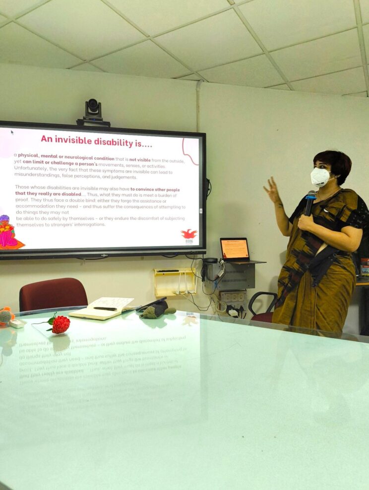 Srinidhi Raghavan standing in front of a TV projecting a presentation showing what an invisible disability is. She has a white face mask on and she is wearing a gold patterned saree with a black blouse. She is speaking into a mic.