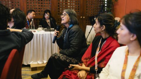 An audience member, Dr. Renu, at the Deconstructing Ableism teach-in speaking into a mic while others listen. In the background are language interpreters sitting at a long table.