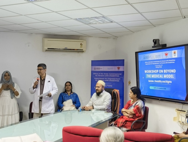 At the head of a conference table are speakers. From the left to right - Dr. Rajesh Patil, Nidhi Goyal, Shri Taha Haaziq, Dr. Shradha Patil. Dr. Rajesh is standing and speaking into a mic. There is a TV on the far right with the event name on it and a standee behind the speakers with the same.