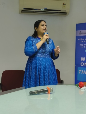 Close up of Nidhi Goyal standing at the head of a table and speaking into a mic. She is wearing a blue long-sleeved kurti.