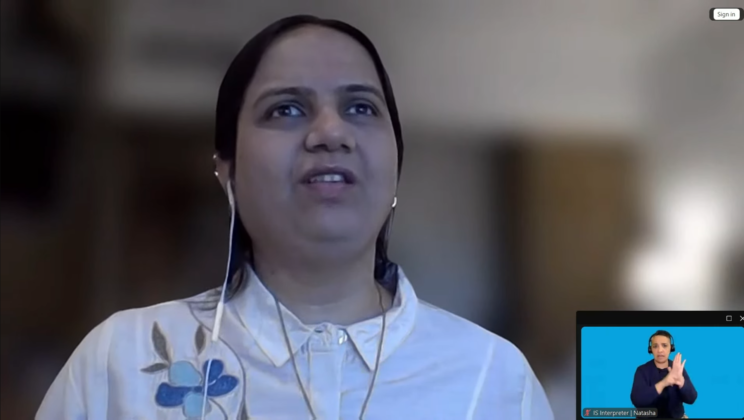 Screenshot of a Zoom window. Nidhi Goyal is speaking. She is wearing a white shirt with a blue floral embroidery on the left side. A window with a sign language interpreter is at the bottom right of the screen.