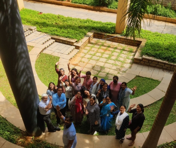 A group photo of men and women taken outdoors from above. They are looking up at the camera, most of them smiling widely. Some of them are waving at the camera, or are posing with peace signs.