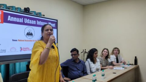 Nidhi Goyal is standing and speaking into a mic. She is wearing a yellow long sleeve kurta. Next to her is a panel of speakers sitting - Prashant Verma, Veena Verma, Stella Elgood Field and Ashleigh Mears. Behind them on a large screen are details of the event: "Annual Udaan Intern Meet" with the Udaan logo below, venue details, and logos of the organisers, partially blocked.