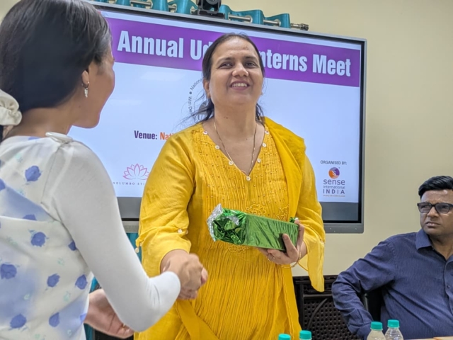 A close up of Nidhi receiving a gift wrapped in metallic green paper and shaking hands with the person handing it to her.
