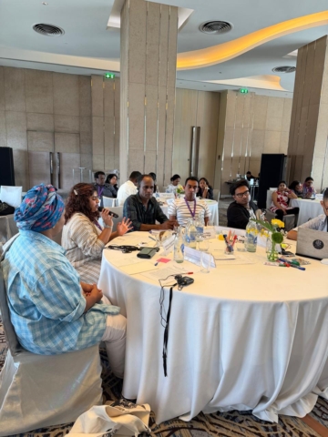 A group of 6 participants at a round table with some other groups visible in the background. One of the participants has a mic and is speaking from her seat.