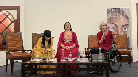 From left to right, Shamim Salim, Nidhi Goyal and Janet Price sitting on a stage on chairs (Janet is in her wheelchair). In front of them is a low table with bottles of water. Behind Janet is a standee that says 'DSROI 10 Years' Anniversary.' Janet is speaking into a mic while the other two listen.