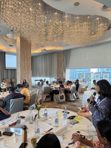 Several round tables with participants, and Nidhi is visible up front. A participant from one of the tables at the back is standing and speaking into the mic.