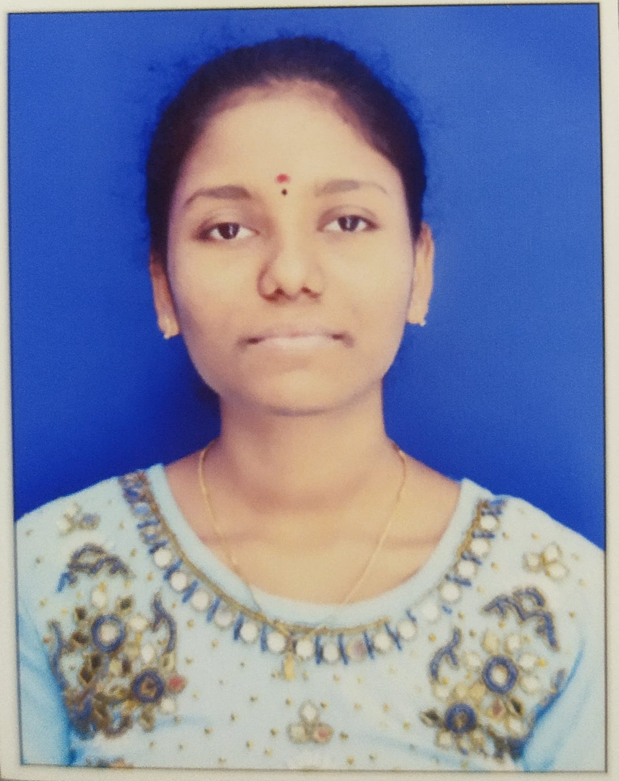 Chandana Balabomma is an Indian woman with her hair tied back. She is sitting in a studio with a blue background, wearing a light kurta with embellishments on the collar and a small red dot on her forehead.