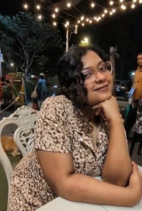 Gayatri has dark curly hair. They are wearing glasses, and a brown and white floral collared top. They are sitting outdoors with a hand on her chin.