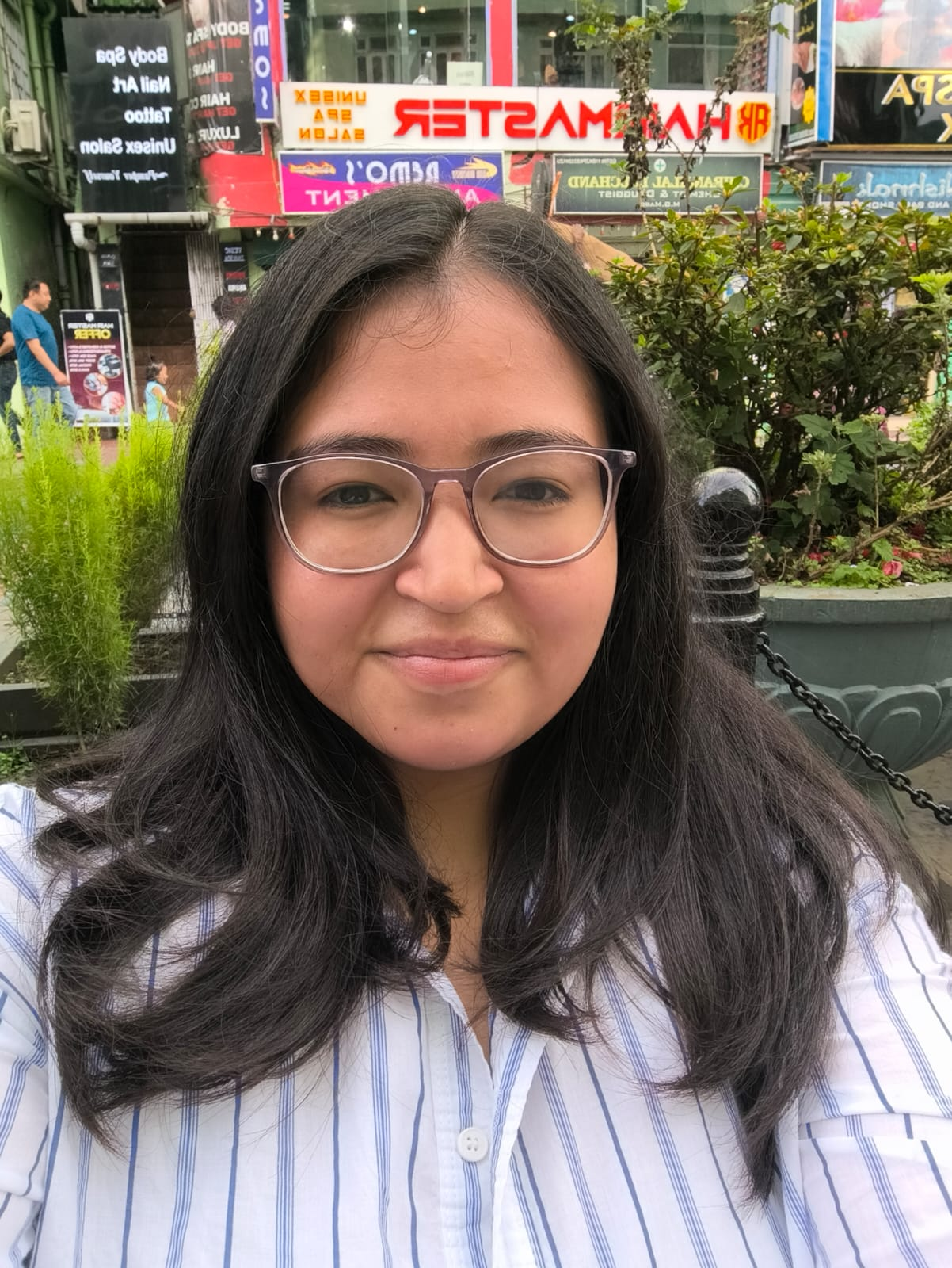 Kaeshikka Chettri is an Indian woman with shoulder-length dark hair. She is wearing glasses and white and blue striped shirt. She is outside - some stores are visible in the background.