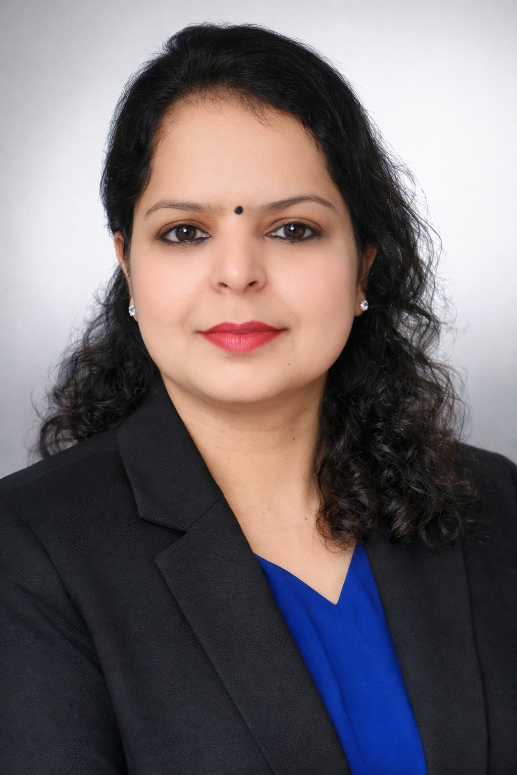 Kanu Mahajan is an Indian woman with curly dark hair. She is wearing a black blazer, sitting in a studio with a white background. She has a small black bindi on her forehead.