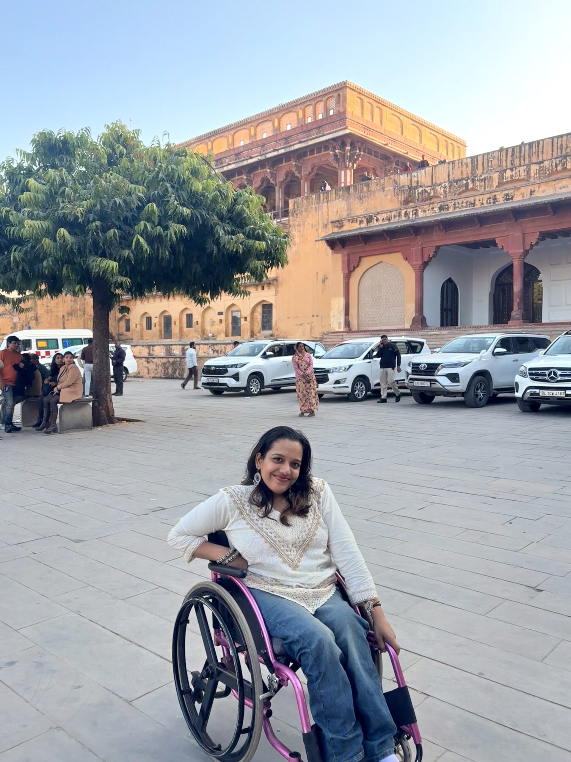 Maithili Gaikwad is an Indian woman who is outdoors, in her wheelchair with pink piping. She has shoulder-length dark hair and is wearing a white kurti with gold embellishments and blue jeans. There are some cars in the background.
