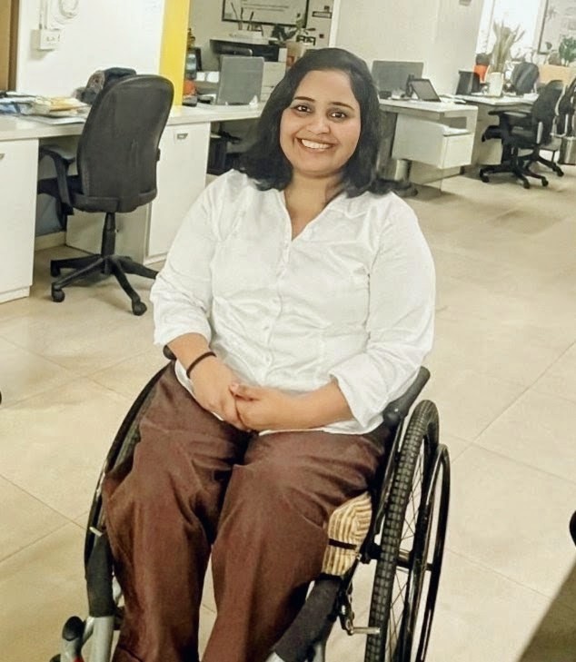 Pratibha Kumari is an Indian woman sitting on her wheelchair inside an office. She has dark, shoulder-length hair, and she is wearing a white shirt and brown pants.