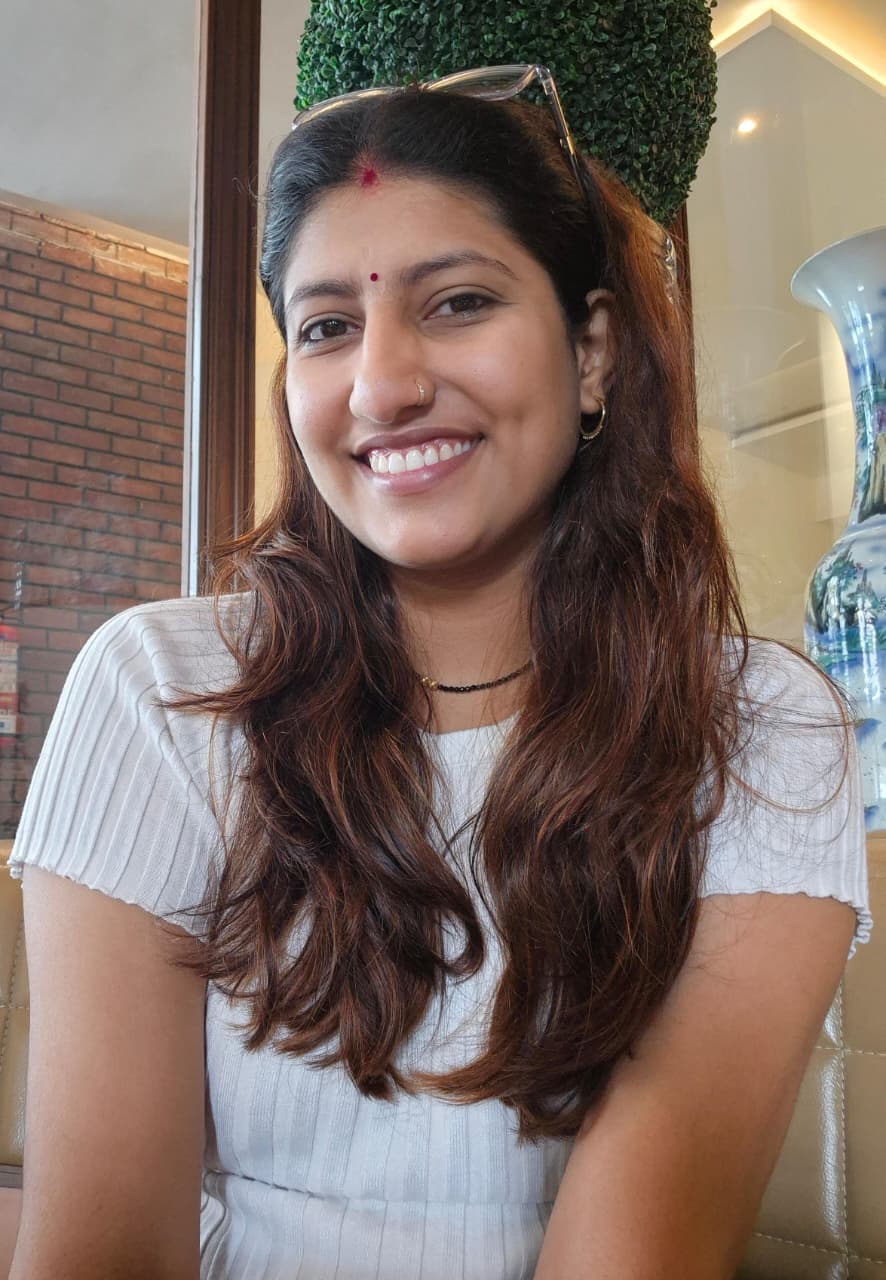 Sapna Jangid is an Indian woman with long hair that is dyed brown. She has propped her glasses up to push her hair back. She is wearing a white short-sleeved top and small hoop earrings. She has some sindoor on her forehead.