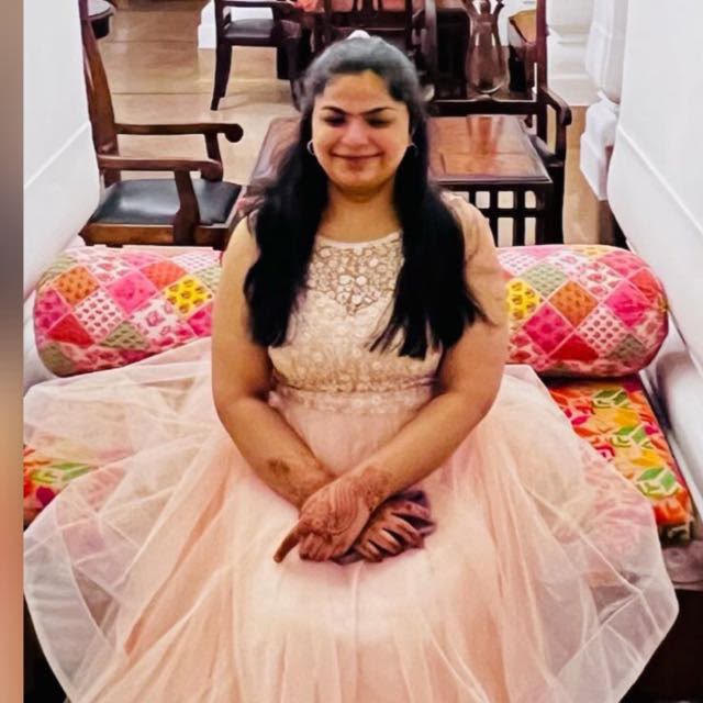 Sara Chawla is an Indian woman sitting on a bright coloured bed. She has long dark hair and is wearing a pink, embellished, sleeveless gown.