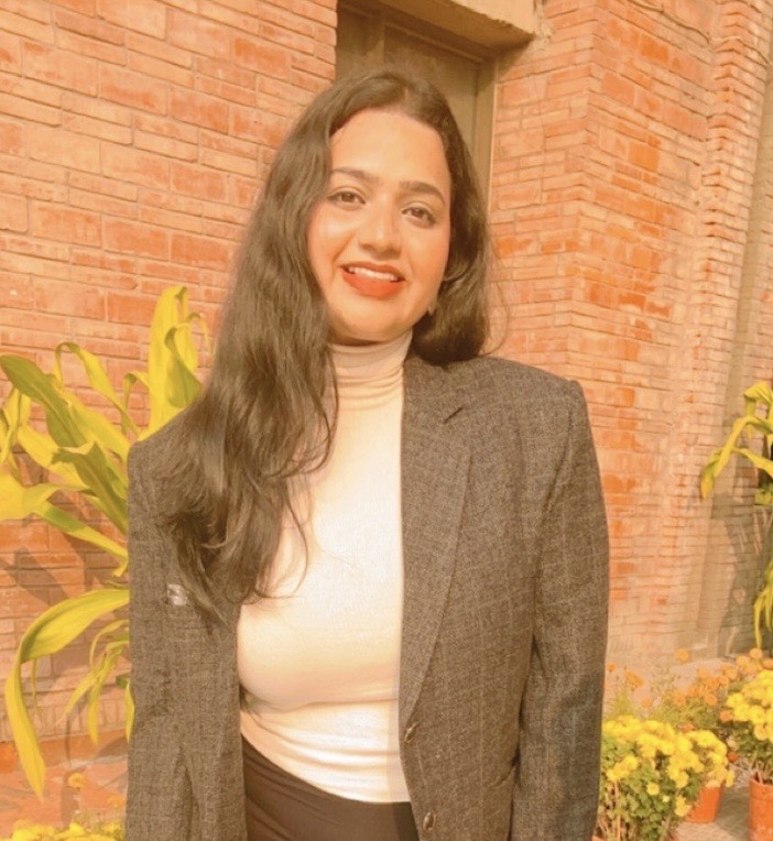 Shafali is an Indian woman with long dark hair. She is standing outdoors with a brick wall in the background. She is wearing a grey blazer over a white turtleneck.