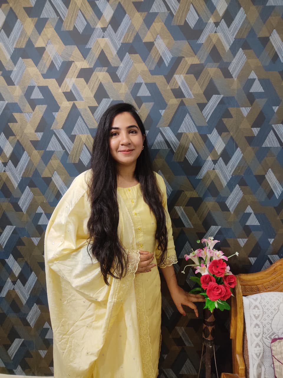 Zainab Madarwala is an Indian woman standing in front of a dark wall. She has long dark hair, and is wearing a yellow Indian suit with a dupatta draped around her arm. There is a vase of flowers next to her.