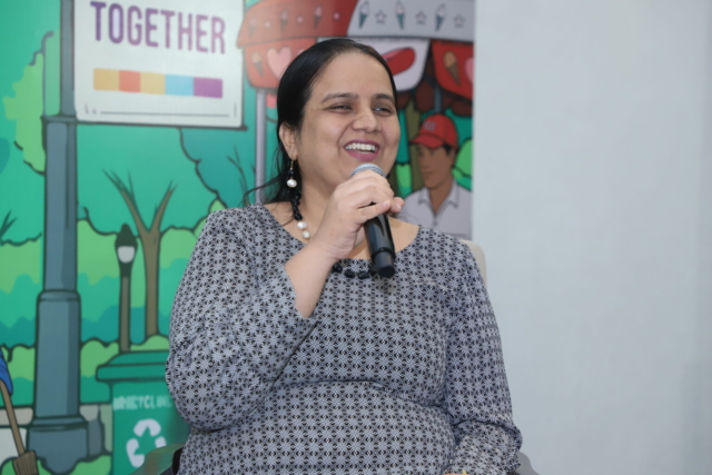 A mid-shot of Nidhi Goyal sitting in front of large banner. She is wearing a black and white patterned long-sleeve kurti and a black and white beaded necklace. She is speaking into a mic, smiling.