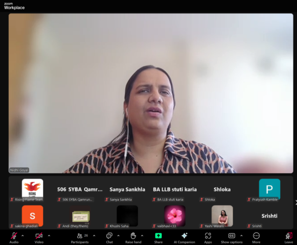 Zoom screenshot with Nidhi Goyal spotlighted, and other participants with their videos and mics off below. She is wearing a brown and black patterned collar shirt and her background is blurred.