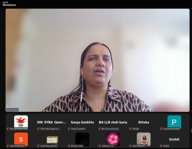 Zoom screenshot with Nidhi Goyal spotlighted, and other participants with their videos and mics off below. She is wearing a brown and black patterned collar shirt and her background is blurred.