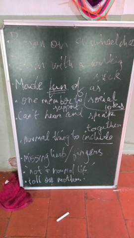 A blackboard leaning against a wall with some points on it. They read: "Person on a wheelchair"; "Person with a walking stick"; "Made fun of as social jokes"; "One member to support"; "Can't hear and speak together"; "Normal thing to include"; "Missing limb/fingers"; 'Not a normal life'; "Toll on mothers"