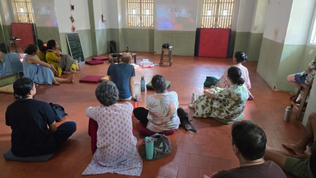 The participants of the workshop, some sitting on the floor, some on chairs and some standing, watching a video of Nidhi Goyal, projected on the opposite wall.