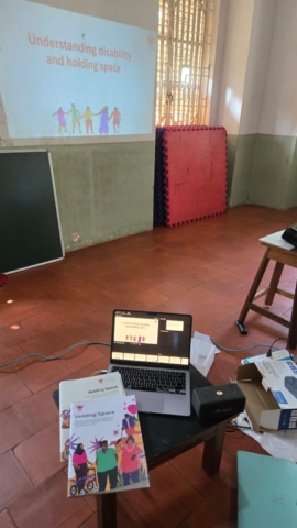 There is a presentation projected on a wall which is titled "Understanding disability and holding space." In front of it is a small table with a laptop and 2 manuals on it - Holding Space Parts 1 and 2.