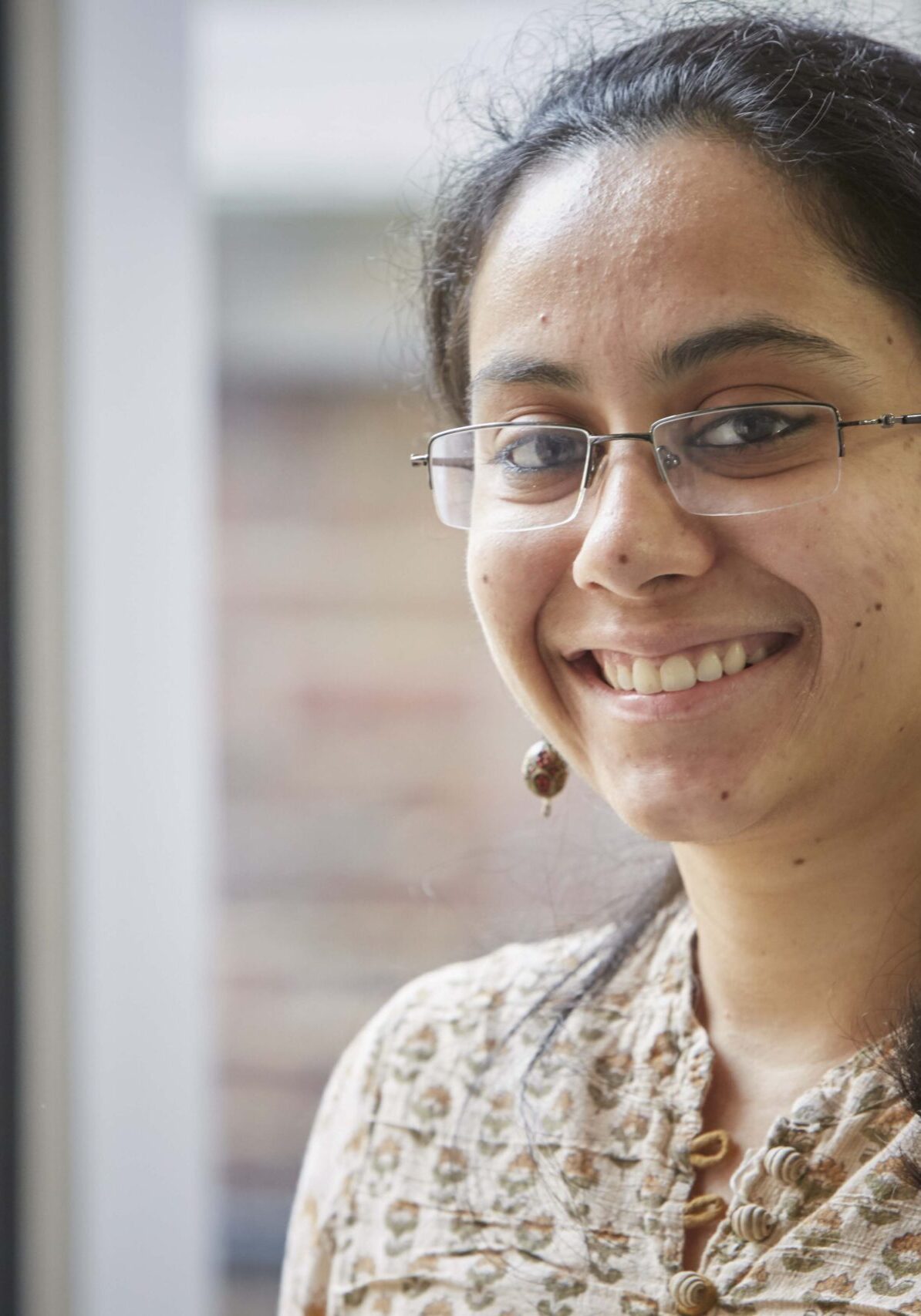 Image of Gayatri smiling by a window, wearing glasses and a patterned kurta.